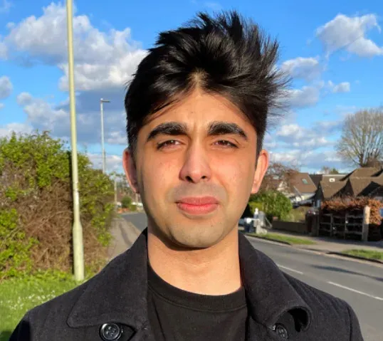 Man with dark hair stands on a suburban street under a blue sky with clouds, wearing a black coat.