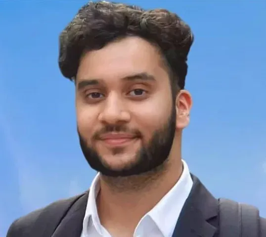 Smiling man with curly hair and beard, wearing a suit and backpack, set against a clear blue sky.